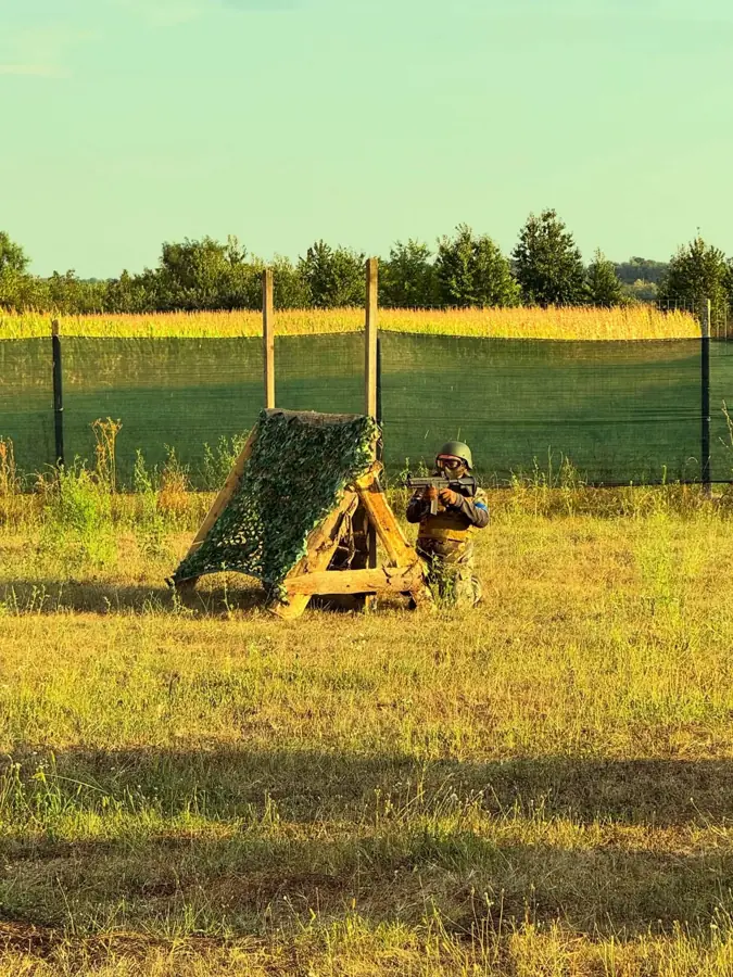 Adăpost din lemn acoperit cu plasă de camuflaj, cu un jucător lângă el